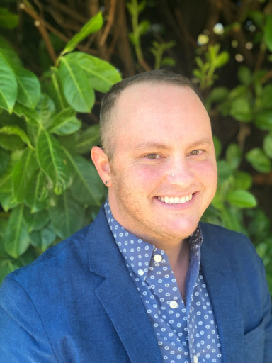 Photo of a smiling person wearing a patterned shirt and blue jacket in front of green foliage